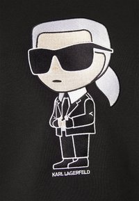 Černá látka obsahuje vyšívanou kreslenou postavičku v obleku s kravatou, s bílými vlasy a velkými černými slunečními brýlemi; text zní "KARL LAGERFELD."