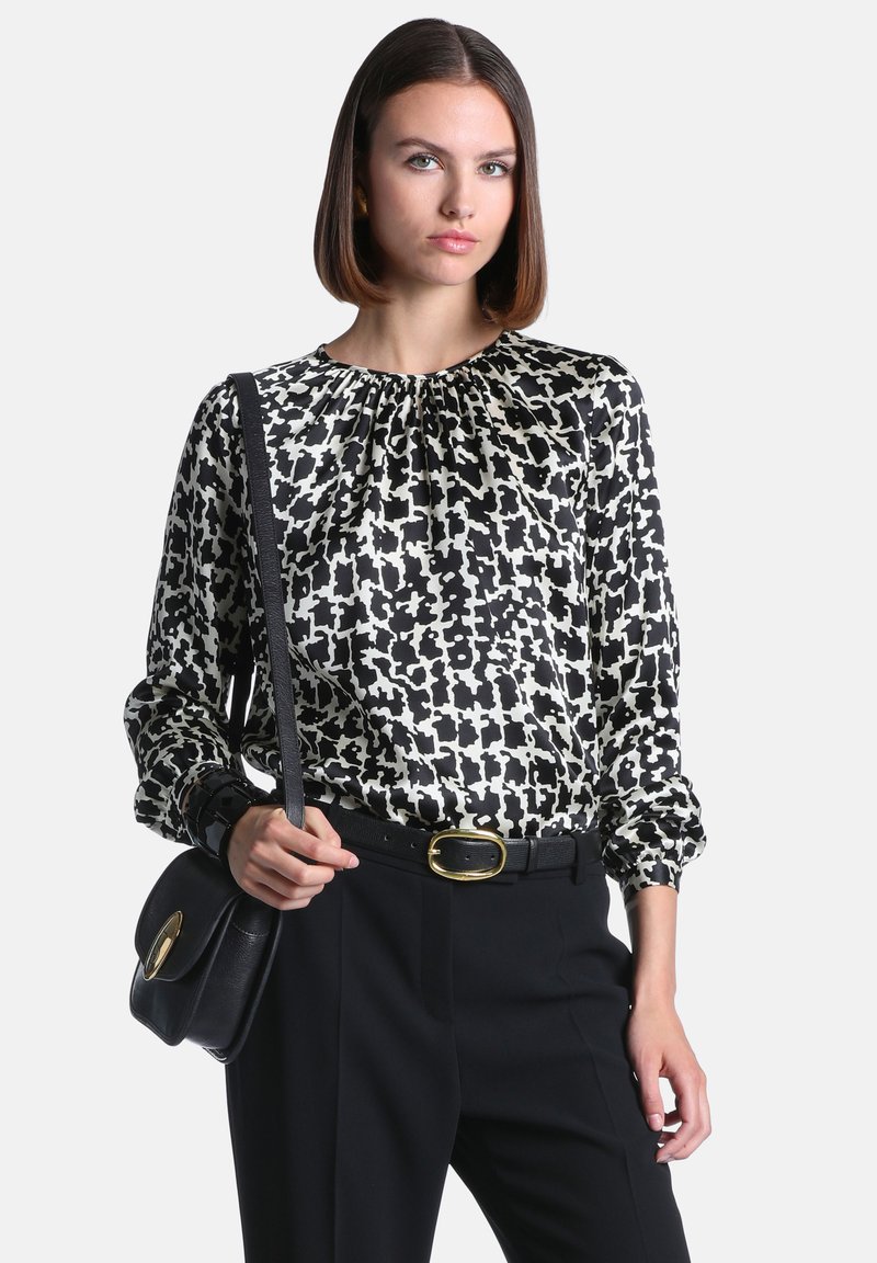 Femme portant un chemisier à imprimé abstrait noir et blanc, un pantalon noir, une ceinture noire avec une boucle dorée et un sac à épaule noir.