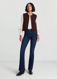 Veste marron sans manches portée sur un chemisier blanc avec des accents en dentelle ; associée à un jean évasé bleu foncé et des bottines noires. Fond uni.