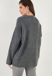 Pulover gri tricotat cu un croi lejer, mansete striate și guler rotund, cu un model texturat și o aventă ușor mai lungă în spate.