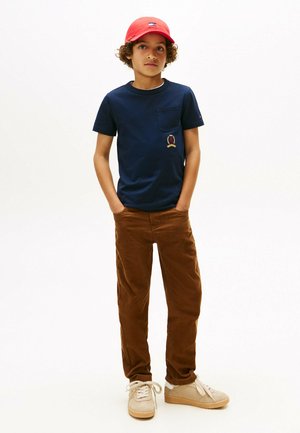 T-shirt en coton bleu marine avec une poche poitrine et un motif brodé, associé à un pantalon en velours côtelé marron et des baskets beiges. Casquette rouge sur la tête.