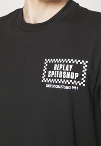 Černé bavlněné tričko s bílým textem "REPLAY SPEEDSHOP" a kostkovaným rámečkem s textem "BIKER SPECIALIST SINCE 1981" pod ním. Krátké rukávy.