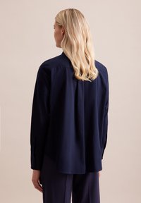 Granatowa koszula z długim rękawem o luźnym kroju, z plisą z tyłu, z zapinanymi mankietami i gładką fakturą materiału, pokazana od tyłu.