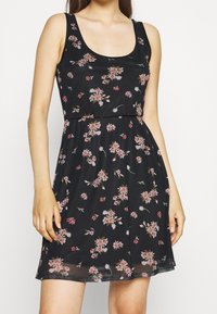 Robe noire à fleurs avec un décolleté arrondi, sans manches, et une jupe fluide, ornée de motifs floraux roses et blancs sur toute la surface.