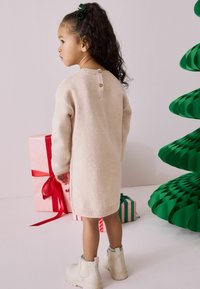 Beige gestricktes Kleid mit langen Ärmeln, das hinten mit Holzknöpfen ausgestattet ist. Kombiniert mit weißen Stiefeln. Im Hintergrund sind Geschenkboxen und ein grüner Papiertree zu sehen.