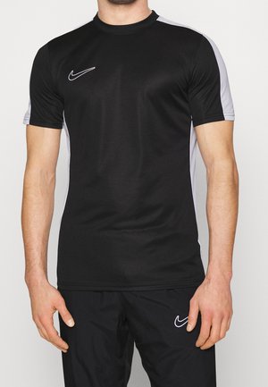 Mann trägt schwarze Nike-Sportbekleidung mit grauen Seitenpaneelen am Shirt und weißen Nike-Logos auf Shirt und Hose, steht vor einem weißen Hintergrund.