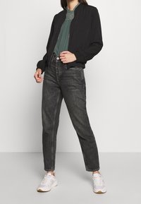 Czarna kurtka bombowiec, zielony top z haftowanymi akcentami, szare dżinsy z wysokim stanem i białe trampki. Strój ma casualowy styl.