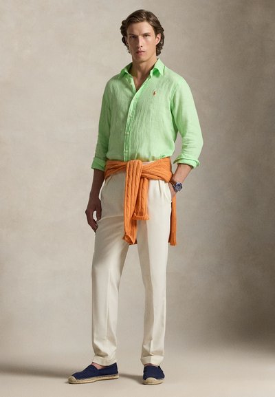 Ανοιχτό πράσινο λινό πουκάμισο, λευκό παντελόνι, πορτοκαλί πλεκτό πουλόβερ δεμένο στη μέση, μπλε εσπαντρίγιες, με ρολόι στον αριστερό καρπό.