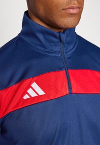 Marineblå idretts half-zip topp med en rød horisontal stripe. Har en hvit logo på stripen og en glatt, teksturert krage.