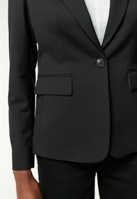 Černý blazer se zapínáním na jeden knoflík, s klopami a bočními kapsami. Vyroben z hladké tkaniny s přiléhavým střihem.