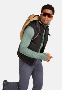 Gilet isolante nero sopra una maglietta a maniche lunghe verde chiaro, pantaloni grigi e occhiali da sole. Uno zaino beige è appoggiato su una spalla.