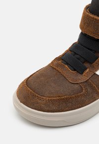 Braune Wildleder-Sneaker mit strukturiertem Obermaterial, schwarzen Textil-Schnürsenkeln und einer weißen Gummi-Sohle. Niedrig geschnittenes Design mit Polsterung um den Knöchel.