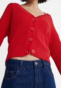 Roter gerippter Strickcardigan mit V-Ausschnitt, fünf großen Knöpfen und verkürzter Passform; kombiniert mit hoch taillierten dunkelblauen Jeans.