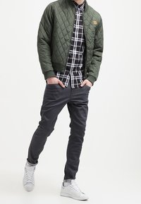 En person står, iklädd en grön vadderad jacka över en rutig skjorta, grå byxor och vita sneakers, framför en enkel ljus bakgrund.