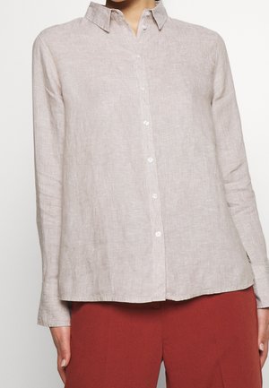 Kobieta ubrana w beżową koszulę na guziki z długim rękawem i kołnierzykiem oraz rdzawobrązowe spodnie, stojąca na jednolitym tle.