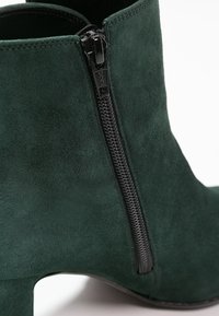 Zelená semišová kotníková bota s bočním zipem a zúženým podpatkem. Materiál má měkkou strukturu a zip je černý s elegantním designem.