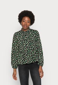 Blusa peplum preta com manchas verdes e creme, apresentando um decote redondo e mangas compridas com punhos franzidos. Textura de tecido suave.