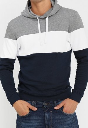 Mand iført en hættetrøje i grå, hvid og mørkeblå farver med hvide snore, hænderne i lommerne, kombineret med blå jeans.