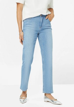 Femme portant un jean bleu clair droit, un haut blanc et des ballerines argentées pointues, une main dans la poche et un bras détendu.