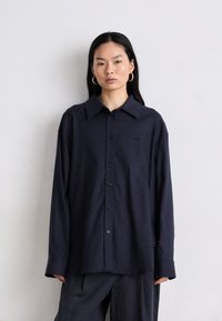 Camisa de manga larga azul marino oscuro con cuello puntiagudo, botones en la parte delantera y un corte holgado. Tejido liviano con textura mínima y un pequeño detalle de logo.