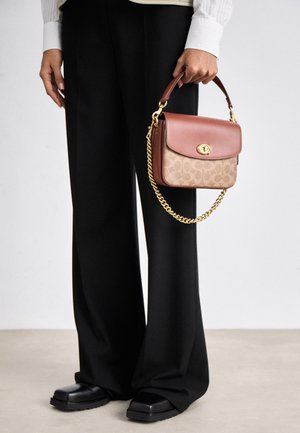 Person trägt schwarze Hosen mit weitem Bein und schwarze Schuhe, hält eine kleine braune und beige Handtasche mit einem goldenen Kettenhenkel.