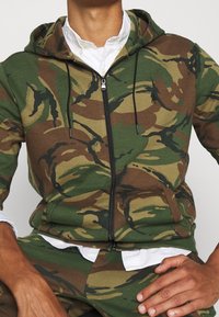 Sudadera con capucha y cremallera en patrón de camuflaje con tonos verdes, marrones y negros; cuenta con bolsillos y un cordón para un ajuste ajustable.