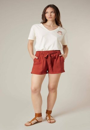 Femme portant un t-shirt blanc à col en V avec des détails floraux, un short noué de couleur rouille, et des sandales marron à bride autour de la cheville, debout les mains dans les poches.