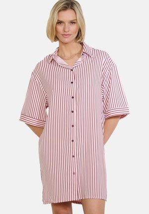 Kobieta ubrana w luźną, do kolan, białą sukienkę zapinaną na guziki z cienkimi pionowymi czerwonymi paskami oraz złoty łańcuszek, stojąca z rękami za plecami.