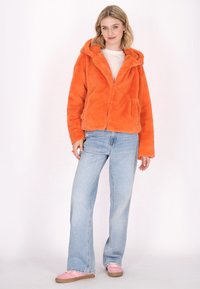 Giacca in finta pelliccia arancione con cappuccio, tasche anteriori e vestibilità rilassata, abbinata a jeans azzurri a gamba larga e sneakers rosa.