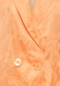 Kahekordse nööbiliistuga virsiku tooniline bleiser, millel on peen lilleline jakardkude ja kaks samas stiilis ümarat nööpi vöö lähedal.