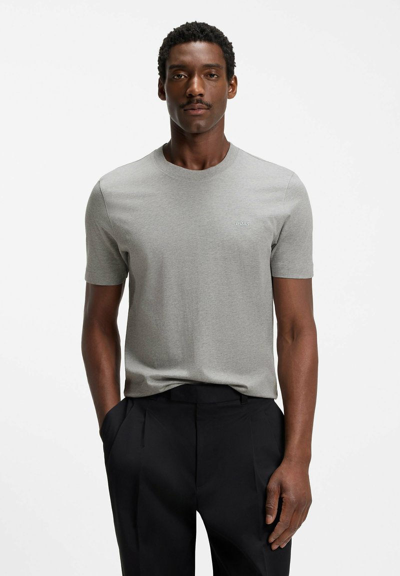 Homme aux cheveux courts et bouclés, portant une chemise à manches courtes gris clair et un pantalon noir, debout devant un fond blanc.
