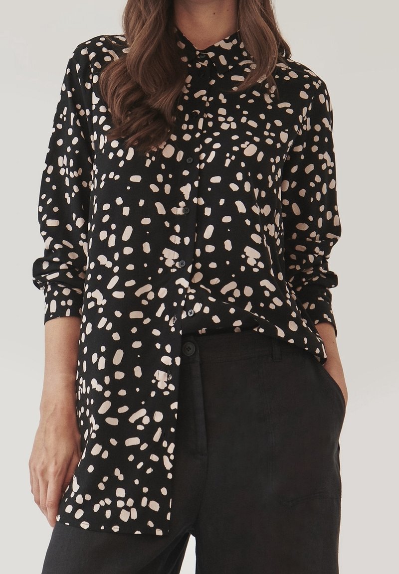 Femme portant une chemise noire boutonnée avec des taches abstraites beige, associée à un pantalon noir, main dans la poche, sur fond clair.