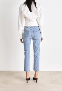 Kvinna iklädd ljusblå jeans, en vit långärmad tröja och svarta klackskor, stående vänd bort på en slät golv och vit väggbakgrund.