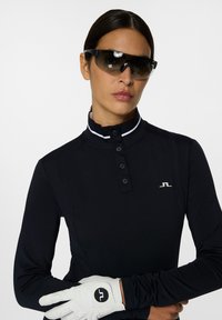 Schwarzes Langarm-Polohemd mit Stehkragen und drei Knöpfen, mit weißen Akzenten. Weißer Golfhandschuh an der rechten Hand getragen. Dunkle Sonnenbrille.