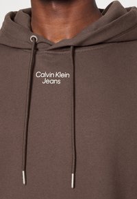 Sudadera marrón hecha de un tejido suave, con un cordón ajustable con puntas de metal y un logo blanco de "Calvin Klein Jeans" en el pecho.