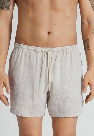 Hombre con shorts de lino beige claro con cordón, mostrando abdomen desnudo y brazos tatuados sobre fondo liso.