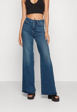 Kobieta w wysokich, szerokich niebieskich dżinsach, czarnym krótki topie i czarnych sandałach na platformie na jednolitym szarym tle.