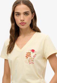 Gul bomulds T-shirt med V-hals, prydet med farverigt broderi af et pagode, blomster og ordet "Sydney" på venstre side.
