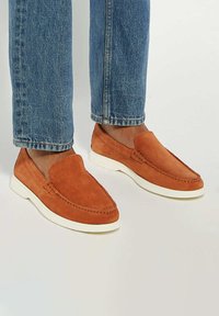 Suede slip-on shoes in orange with a white rubber sole, featuring a moccasin design and stitching detail on the toe.