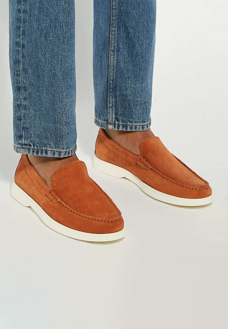 Suede slip-on shoes in orange with a white rubber sole, featuring a moccasin design and stitching detail on the toe.
