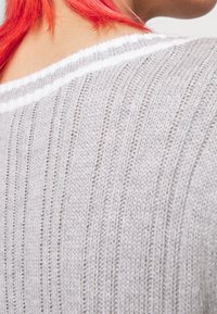 Person med livligt röd hår som bär en ljusgrå ribbad stickad tröja med en vit randad halsringning, visad från axeln och nackområdet.