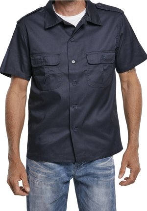 Homme portant une chemise bleu marine à manches courtes avec deux poches frontales et un jean en denim bleu clair, debout les bras détendus.