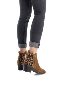 Stivali alla caviglia in suede marrone con pannello laterale in stampa leopardata, chiusura con zip e tacco impilato. Jeans scuri visibili con risvolto.