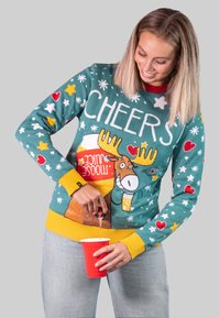 Weihnachtspullover mit grünem Grundton, gelben Akzenten und festlichen Mustern, darunter Elche, Herzen und Sterne. Rotes Getränkebecher in der Hand.