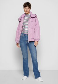 Chaqueta acolchada rosa con capucha, con un patrón acolchado. Llevada sobre una camiseta de manga larga a rayas y unos vaqueros acampanados azules, completada con zapatillas blancas.