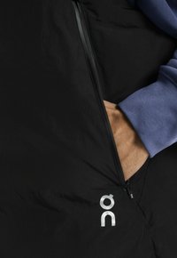 Chaqueta de plumón negra con un bolsillo con cremallera, que presenta un logo plateado. El cuello es de un punto azul claro, ofreciendo un contraste en textura y color.