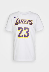 T-shirt blanc à manches courtes avec le logo des Los Angeles Lakers en violet et jaune et le numéro 23 sur le devant, ainsi qu’un petit swoosh Nike violet.