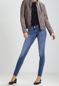 Chaqueta de cuero marrón con escote redondo y diseño ajustado, combinada con jeans ajustados azules y tacones negros. Textura suave en toda la prenda.