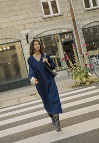 Mornarsko jeansa obleka z dolgimi rokavi in V-izrezom, parjena s črnimi škornji do kolen. Črna torba čez ramo in urbana okolica v ozadju.