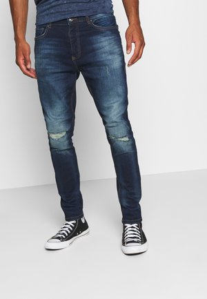 Homme portant un jean skinny bleu foncé usé et des baskets montantes noires, debout devant un fond blanc uni.